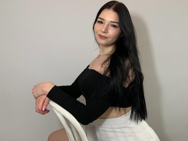 AvaCandy with long black hair, black off-shoulder top, white skirt, sitting on white chair, smiling, looking at camera, hands on chair, pale skin, cute, subtle makeup.