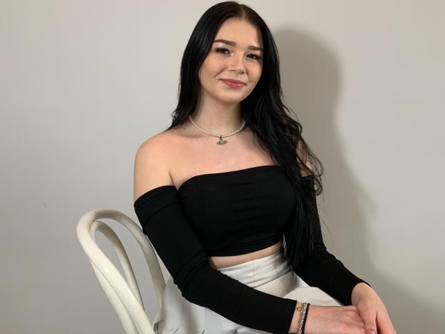 AvaCandy with long black hair, smiling, wearing a black off-shoulder top, white skirt, necklace, and bracelet, sitting on a white chair. Boobs visible.