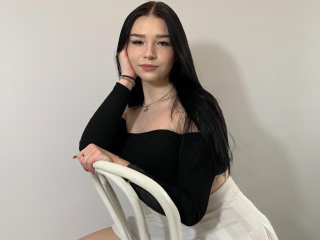 AvaCandy with long black hair, wearing a black off-shoulder top and white skirt, sitting on a white chair, hand on cheek, smiling, looking at camera.