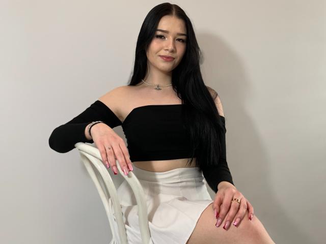 AvaCandy with long black hair, wearing a black off-shoulder top and white skirt, sitting on a white chair, smiling slightly, with pink nails and a gold ring.