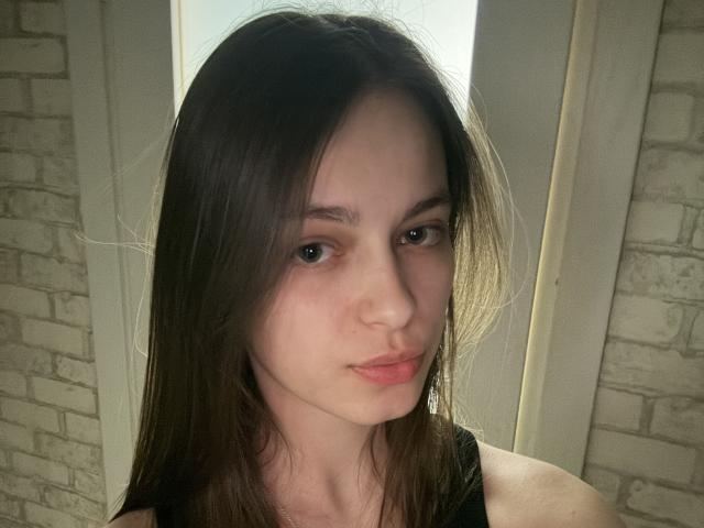 Raichell has pale skin, brown eyes, and long brown hair. She's wearing a black top with a serious, slightly pouty expression. Her small nose and subtle lips are visible.