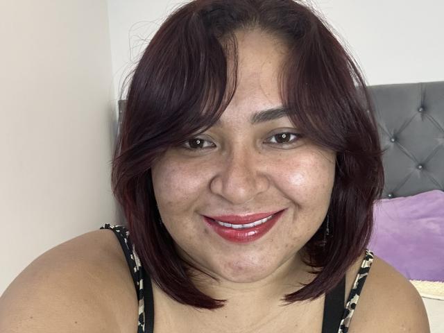 KarlaWild has dark red, shoulder-length hair, brown eyes, and red lipstick. She's smiling with her tits barely visible in a black and white patterned top.