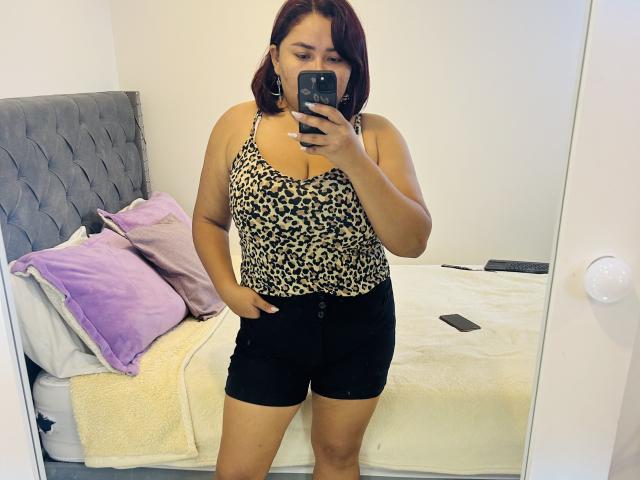 KarlaWild with medium brown skin, red hair, wearing leopard print top, black shorts, holding phone, showing her boobs in mirror selfie, bed with purple pillow and white blanket.