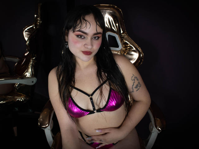 Laila Durmont mit nassen, schwarzen Haaren, einem pinken, metallischen BH und Höschen, einem Tattoo am rechten Arm, der Hand auf dem Bauch, sitzend auf einem goldenen Stuhl, dunklem Hintergrund, blickt mit roten Lippen in die Kamera..