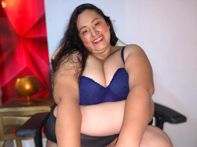 TistMature, une femme corpulente avec de longs cheveux noirs, souriante, portant un soutien-gorge en dentelle bleu, exposant de grandes seins, et des sous-vêtements noirs, assise sur une chaise..