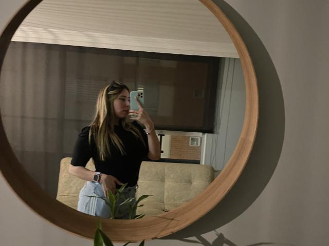 JennyLawry with long blonde hair, black top, and light blue jeans, taking a mirror selfie with a phone, wooden circular mirror, and beige couch in the background.