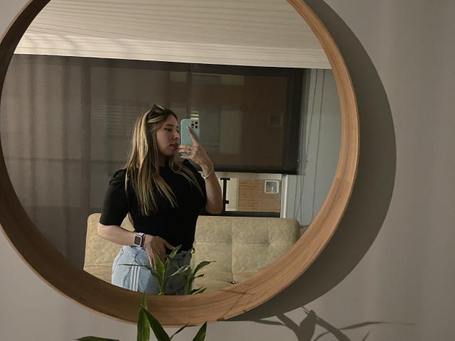 JennyLawry with long blonde hair, black shirt, and light blue jeans, taking a selfie in a circular wooden mirror, showing her ass and wristwatch.