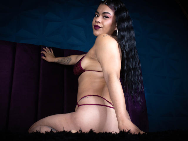 MiahbelleE in a maroon thong and bra, sitting on a dark purple velvet couch, showing her ass and small tits, with long black hair and dark lipstick.