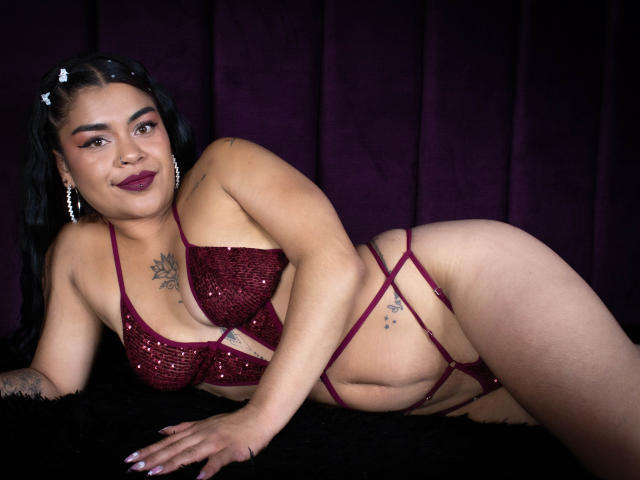 MiahbelleE with dark hair, wearing a red sequin bra and strappy thong, lying on black fur, smiling, showing tattoos, large tits, and a curvy, sexy ass.