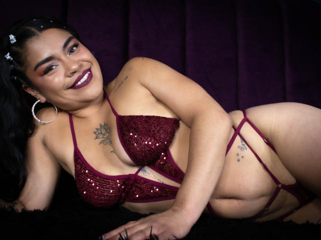 MiahbelleE with dark skin, black hair, wearing a maroon sequined bra and strappy panties, smiling, lying on a black fur, showing tattoos on chest and hip. Boobs, ass, pussy.
