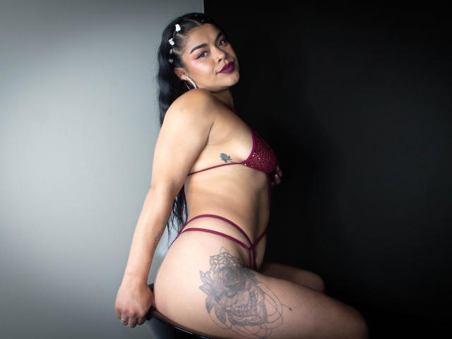 MiahbelleE with long black hair, dark lipstick, and maroon strappy lingerie, showing her ass, tits, and thigh tattoo, sitting on a black stool.