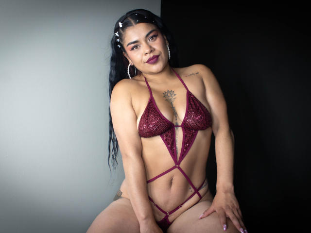 MiahbelleE with dark hair, wearing a sparkly red bikini, showing small tits, a tattoo on her chest, and sitting with her hand on her pussy. She has hoop earrings.