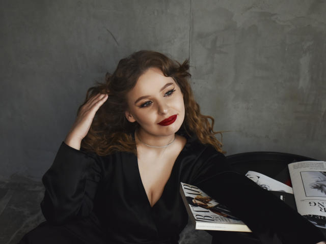 KyleJackson with curly brown hair, red lipstick, black dress, holding a magazine, sitting, hand in hair, grey wall, silver necklace, black chair, book on lap, sexy.