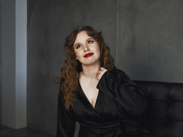 KyleJackson with curly brown hair, red lipstick, and black satin dress, hand on chest, sitting on black leather chair, grey wall behind. Her tits are small, she's cute.