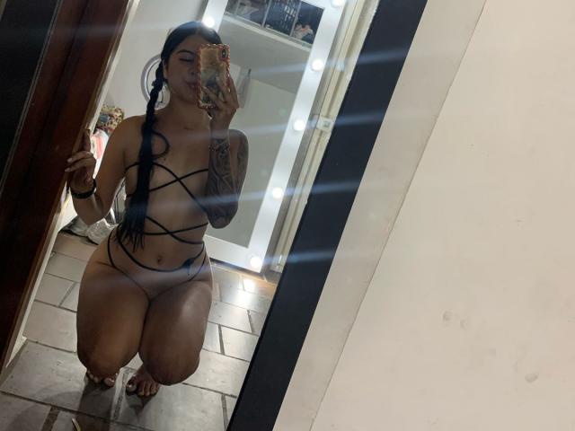 NicoleCollins kneeling in front of a mirror, wearing strappy black lingerie, showing her tits and pussy, with tattoos on her arm, taking a selfie with a phone.