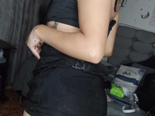 A MaiteX tem uma pequena tatuagem no lado direito, veste uma cropped preta e uma saia preta de cintura alta, com a mão na cintura, mostrando o bumbum..