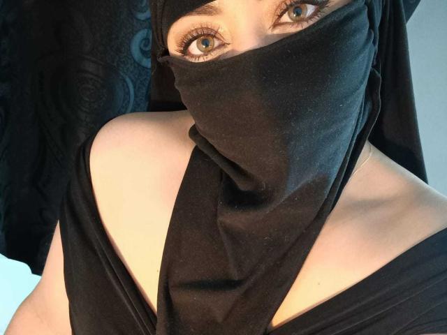 ZahraJalid has blue eyes, wearing a black mask and headscarf, exposing bare shoulders and a black top. Boobs are partially visible. Her face is mostly covered.