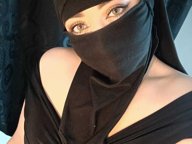 ZahraJalid has blue eyes, wearing a black burqa with a veil, exposing her shoulders and upper chest. Her long eyelashes are visible. Black fabric covers her nose and mouth.
