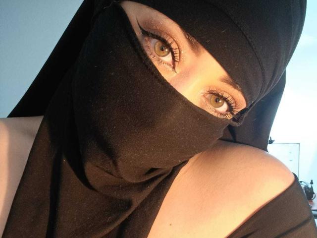 ZahraJalid with a black mask covering her lower face, exposing her golden-green eyes and long eyelashes, wearing a black headscarf, in a warm-lit photo.