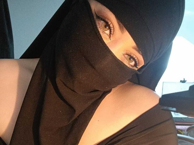 ZahraJalid with yellow eyes, black mask, and black head covering, exposing bare shoulders, wearing black off-shoulder top, holding black glove near face.