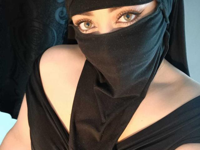 ZahraJalid with blue eyes, wearing a black headscarf and mask, exposing her shoulders and upper chest, in a black top. Her eyelashes are long and slightly curled.