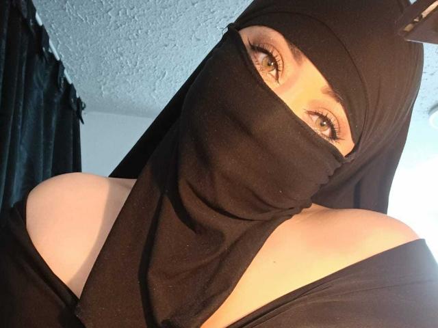 ZahraJalid with black mask covering her mouth and nose, wearing a black headscarf with a small horn, exposing her green eyes and bare shoulders. Her tits are partially visible.