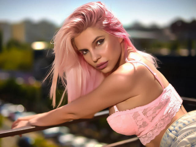 GoddessKaterina, avec ses cheveux roses, son soutien-gorge en dentelle rose et son jean en denim, appuyée sur une rambarde, montrant ses seins, apparaissant sexy avec des yeux séduisants et un léger sourire..