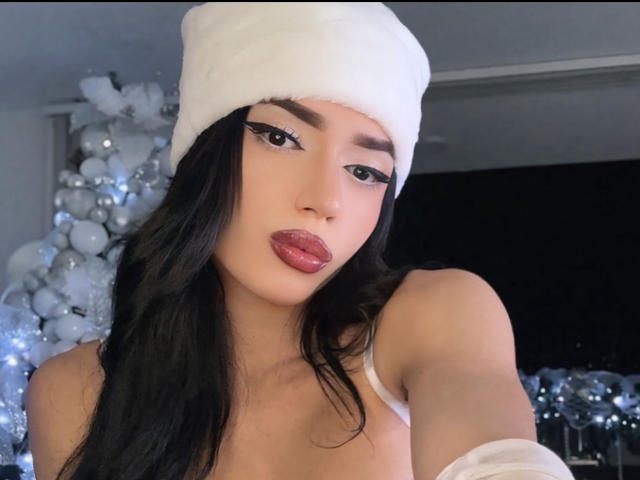 SarayQueen with long black hair, white furry hat, and glossy pink lips, wearing a white top, showing her bare shoulders and upper chest, with prominent eyeliner.