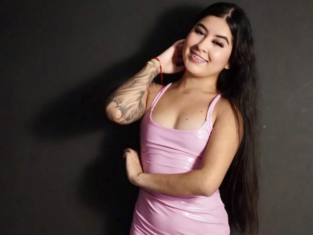 NicoleCollins with long black hair, pink shiny dress, tattooed arm, smiling, hand on hip, pink lipstick, dark background. Boobs, ass, pink dress, tattoo.