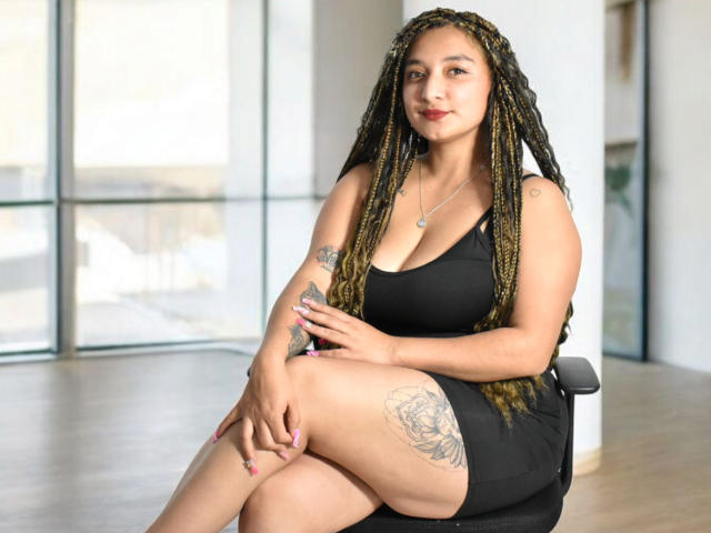FantastykLadies with long braided hair, black dress, showing a thigh tattoo, sitting on a chair, smiling, showing her tits, and crossed legs. She has a necklace and pink nails.