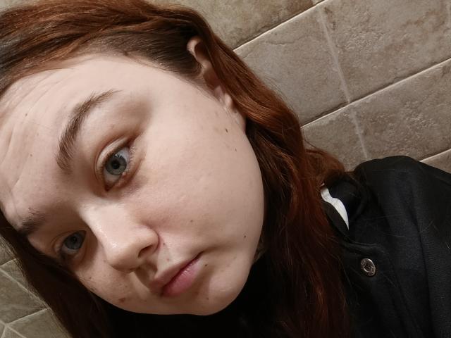 ArielXxl has pale skin, blue eyes, and reddish-brown hair. She's wearing a black shirt, looking slightly off-camera with a neutral expression and a small mole on her nose.