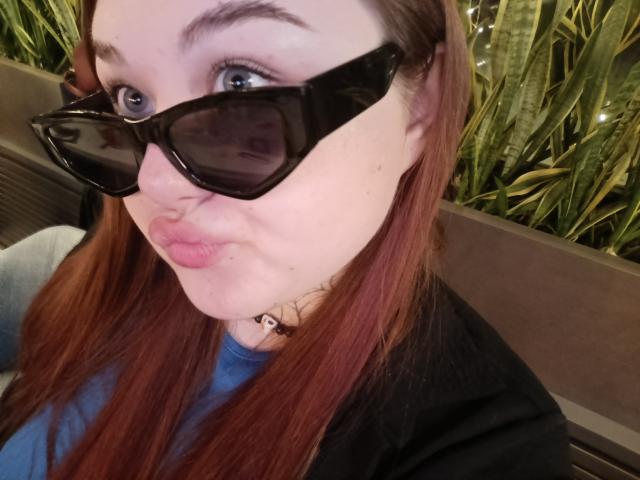 ArielXxl with red hair, puffy pink lips, blue eyes, black cat-eye glasses, black choker, and blue shirt, making a pouty face. Her ass is not visible.