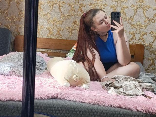 ArielXxl with long red hair, blue sleeveless top, and black choker, sitting on a pink fluffy bed, taking a mirror selfie, showing her face and tits.