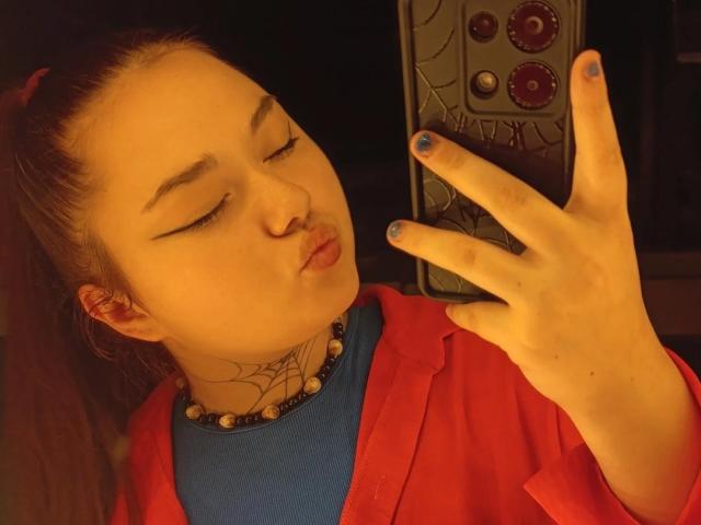 ArielXxl with pouty lips, blue nails, red jacket, and blue shirt, holding phone with spider design, tattoo on neck, brown hair in ponytail, yellow light.