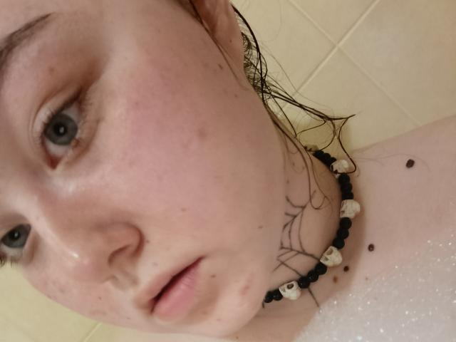 ArielXxl's pale face with freckles, wet brown hair, blue eyes, and black-bead necklace with white shells, showing small moles near the neck, in a foamy bath.