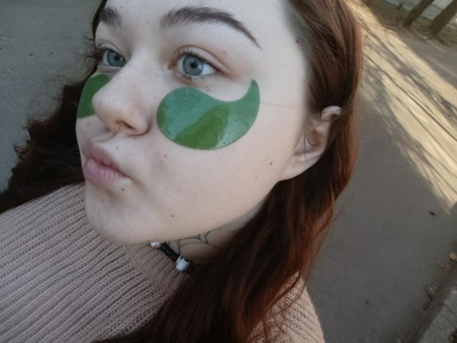 ArielXxl with green paint circles on her cheeks, pale skin, blue eyes, brown wavy hair, pink ribbed shirt, and a necklace, looking pouty in a selfie.