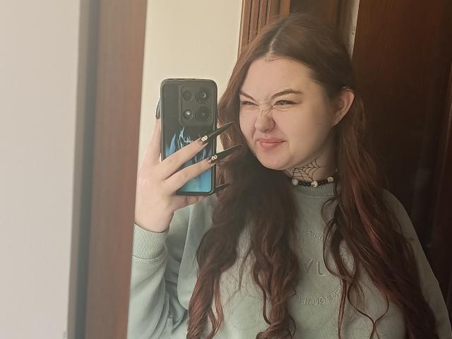 ArielXxl with long, wavy brown hair, wearing a green sweater, taking a selfie in a mirror with a blue phone, smiling, showing off her tits and black nails.