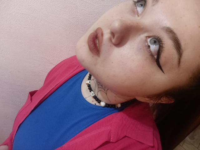 ArielXxl with blue shirt, pink jacket, black choker, and tattoo, staring up with grey eyes, pouty lips, and thick black eyebrows, head tilted back.