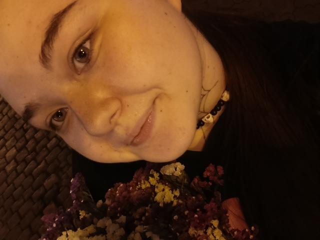 ArielXxl's face with a slight smile, brown eyes, and freckles, holding a bouquet of purple, yellow, and pink flowers, wearing a black top and bead necklace.