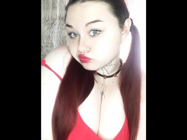 ArielXxl with red pigtails, blue eyes, and red lipstick, wearing a red top that shows her big tits, a black choker, and a tattoo on her chest.