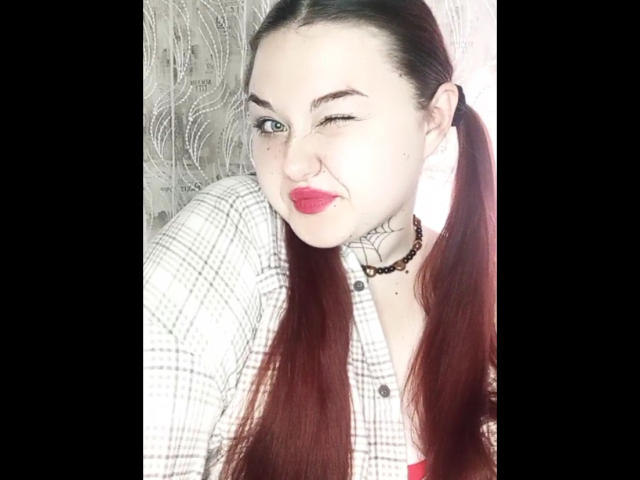ArielXxl with red lipstick, winking, has long red pigtails, white plaid shirt, black choker, and a heart tattoo on her neck. Her tits are small, she's cute.