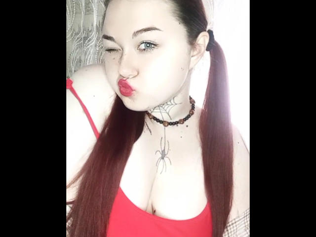 ArielXxl with pigtails, red lips, and a spider tattoo, wearing a red top and black choker, sucking on her finger, showing her tits. Her eyes are blue.