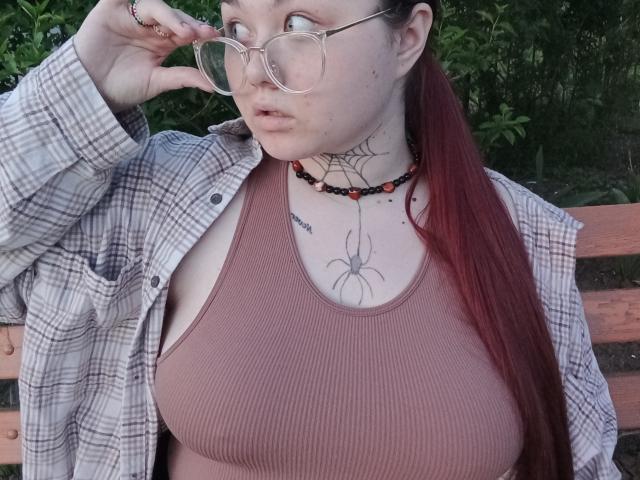 ArielXxl with red hair, glasses, plaid shirt, and pink tank top, showing large tits, spider tattoo, and beaded necklace, sitting on wooden bench.