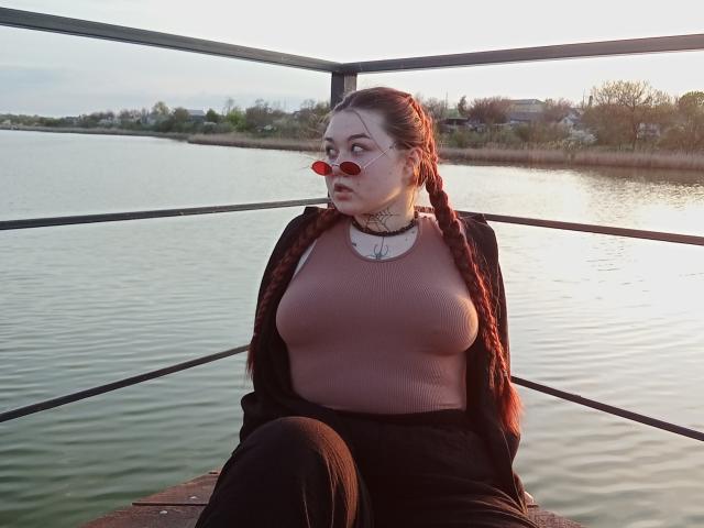 ArielXxl with red braids, pink top, black jacket, black pants, large tits, red heart sunglasses, white necklace, sitting on wooden dock by calm water, metal rail behind.