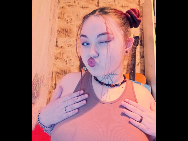 ArielXxl with puffy lips, winking, wearing a brown tank top, hands on chest, tattoos, beaded necklace, ring, and bracelet, hair in a bun, wall behind.