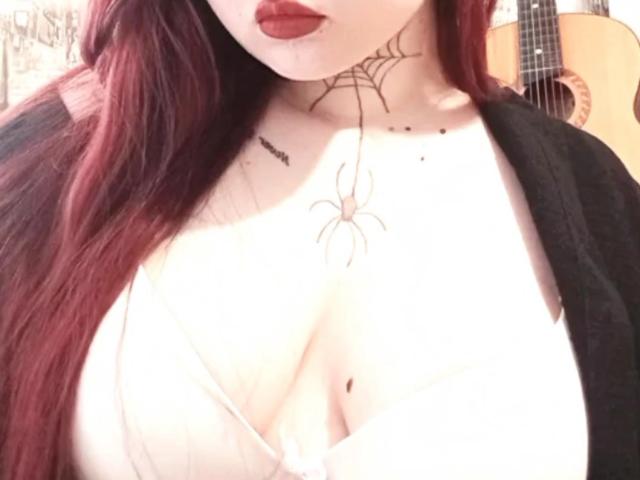 ArielXxl with red hair, pale skin, and red lipstick, showing large boobs with a spider tattoo between, wearing a white top and black cardigan.