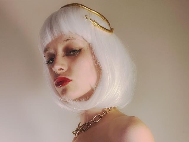 Déesse Divonix avec une coupe de cheveux courte blanche, un halo doré, des lèvres rouges, un trait d'eye-liner prononcé, un collier en chaîne, une peau pâle et une expression sérieuse, mettant en valeur ses seins..