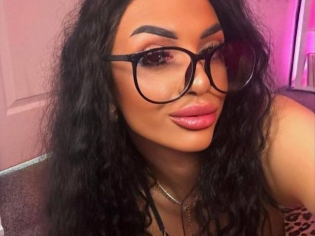 SamiraVeda, con i capelli neri mossi, indossa grandi occhiali neri e rossetto rosa, ha un sorriso carino ed è intenta a mostrare il suo brillante e prosperoso sedere..