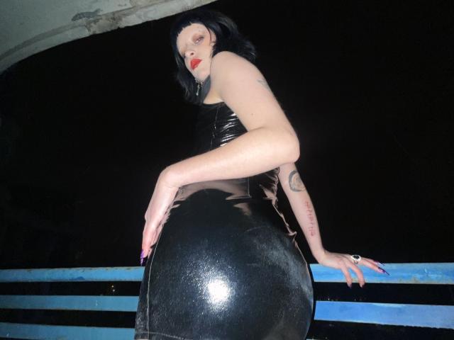 GoddessDivonix with pale skin, black latex dress, red lipstick, and black nails, leaning on blue railing, showing her shiny black ass, tattoos on arm, dark hair, night.