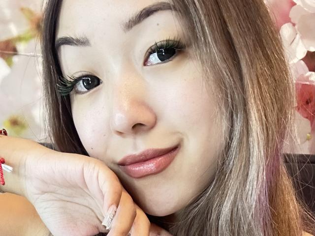 RenyLime with long brown hair, green eyes, and glossy pink lips, smiling slightly with her hand resting on her cheek. She has green eyeliner and a red bracelet.